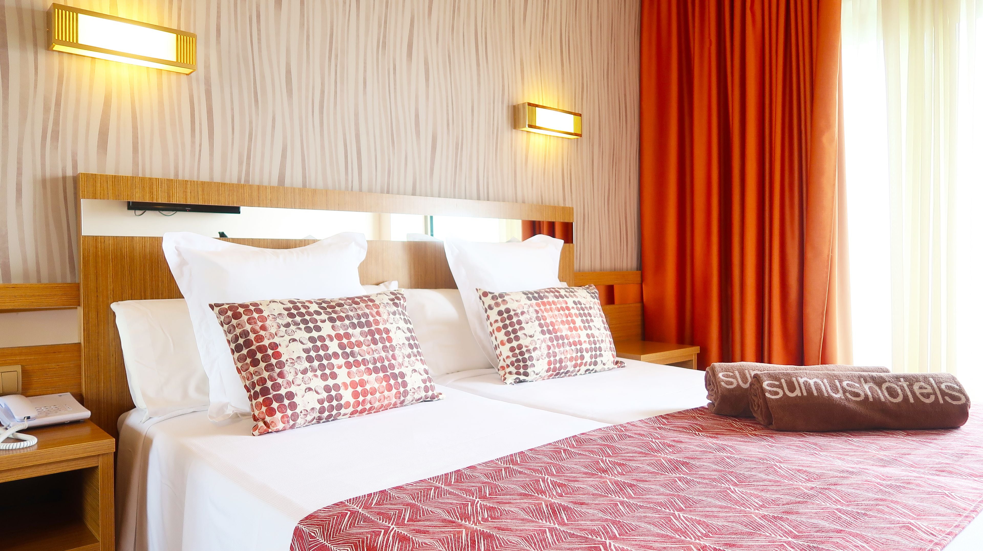 Hotelkamer met een houten hoofdbord, twee bedden met witte lakens, gestreepte kussens, een bedspread met een rood-wit patroon, een bruine handdoek met tekst, een nachtkastje met een telefoon, en twee wandlampen.