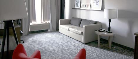 A hotel room with a red chair, a white sofa, a small round table, a lamp, and a framed picture on the wall.
