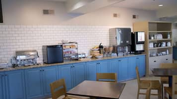 A kitchen with blue cabinets, a granite countertop, a refrigerator, a microwave, and a coffee machine. There are wooden chairs and a table.