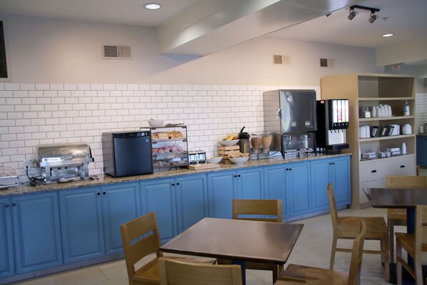 A kitchen with blue cabinets, a granite countertop, a refrigerator, a microwave, and a coffee machine. There are wooden chairs and a table.
