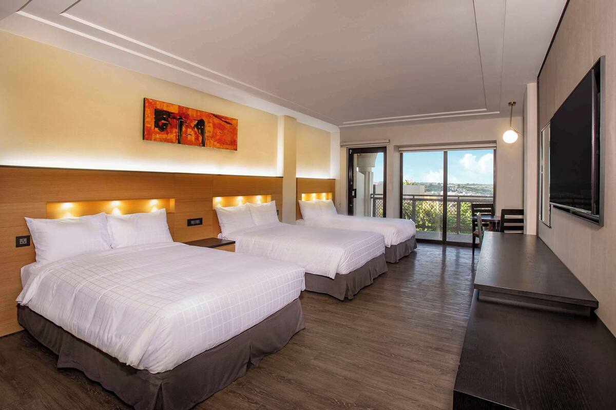 A hotel room with three beds, a large TV, and a balcony with a city view.