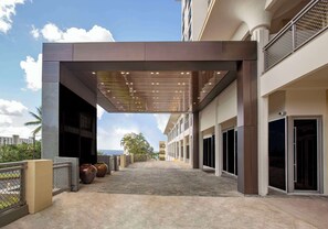 A covered outdoor area with a modern design, featuring a pathway leading to a building entrance, large potted plants, and a view of the ocean. - Bayview Hotel Guam (Tamuning)
