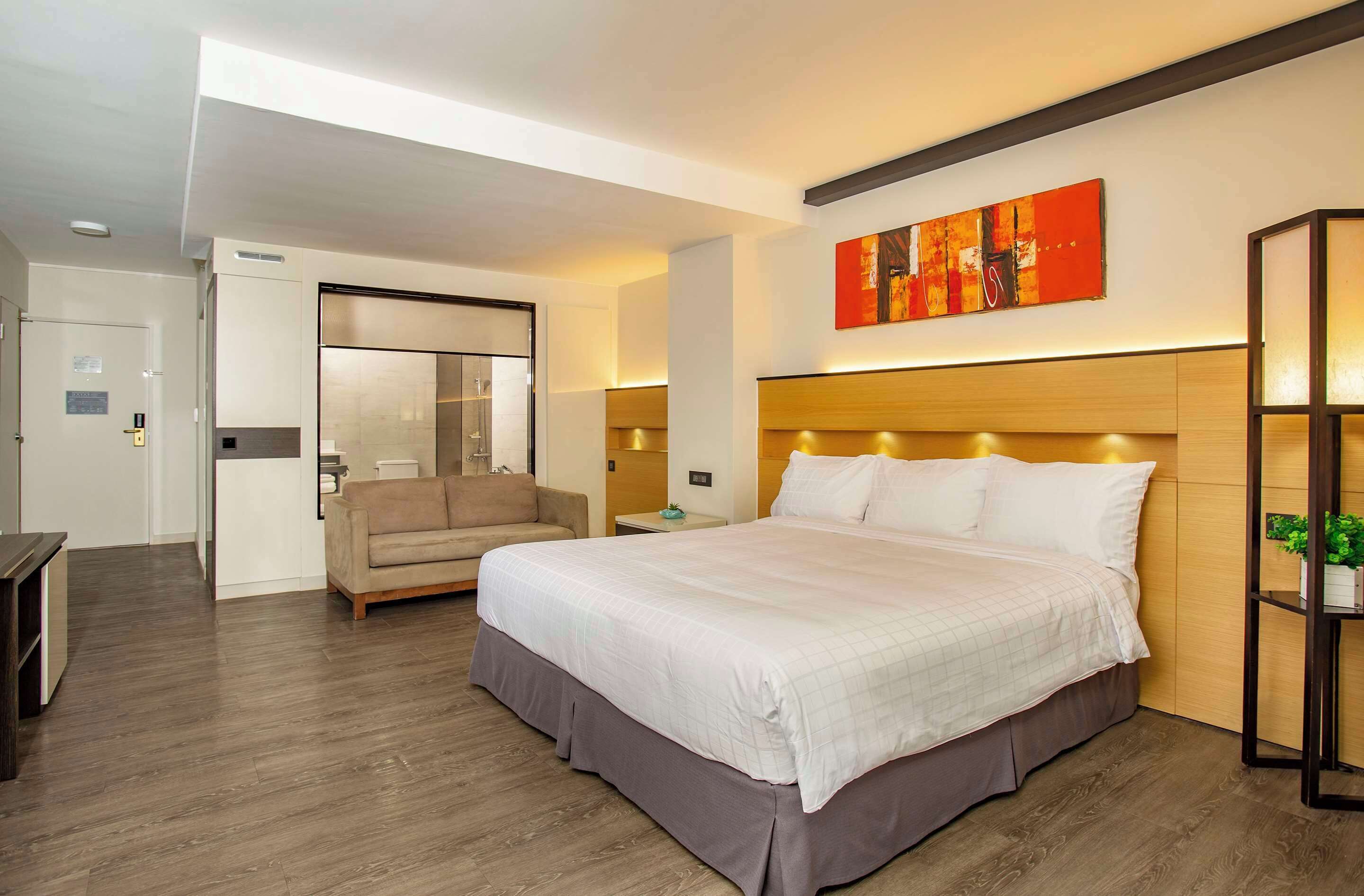 A modern hotel room with a large bed, a sofa, a bedside table, and a wall-mounted artwork.