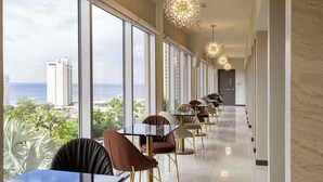 A hotel corridor with tables and chairs, large windows with a view of the ocean and buildings, and decorative ceiling lights. - Bayview Hotel Guam (Tamuning)