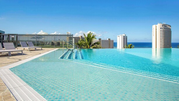 A swimming pool with tiled flooring, lounge chairs, and a view of the ocean and buildings. - Bayview Hotel Guam (Tamuning)