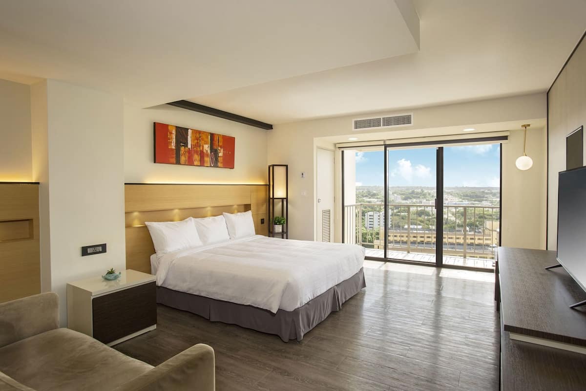 A hotel room with a bed, a desk, a chair, a TV, a painting, and a balcony with a city view.