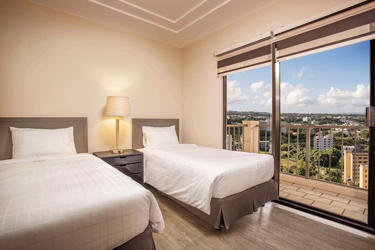A hotel room with two beds, a nightstand with a lamp, and a large window offering a city view.