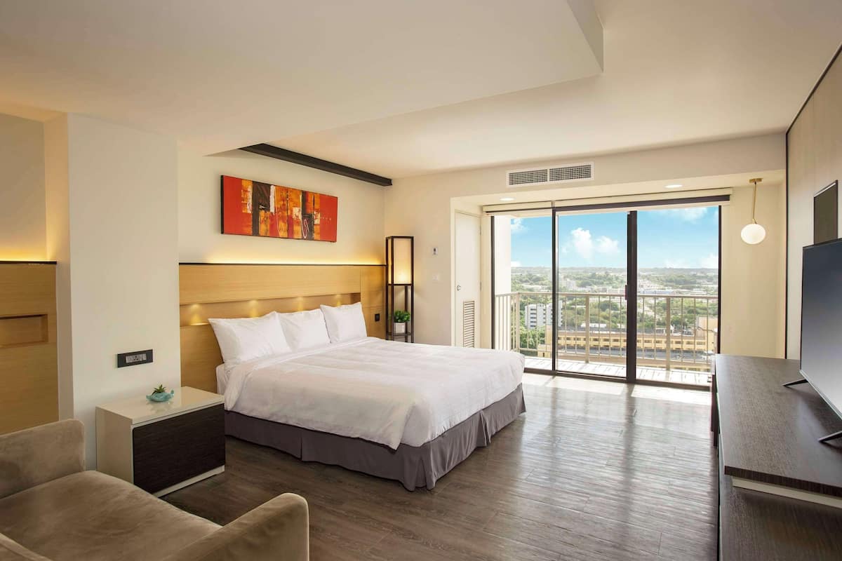 A modern hotel room with a large bed, a TV, a desk, and a balcony with a city view.