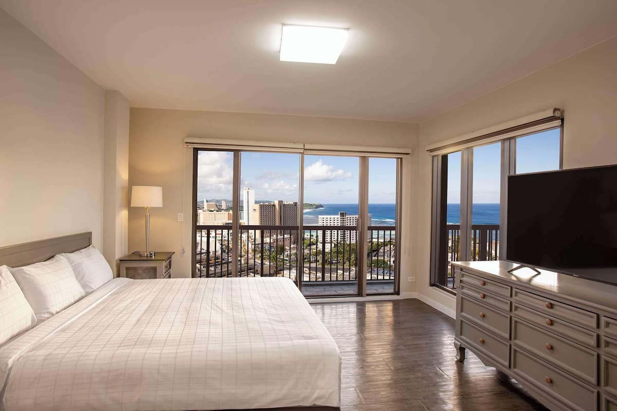 A hotel room with a large bed, a TV, a dresser, and a balcony with a city view.