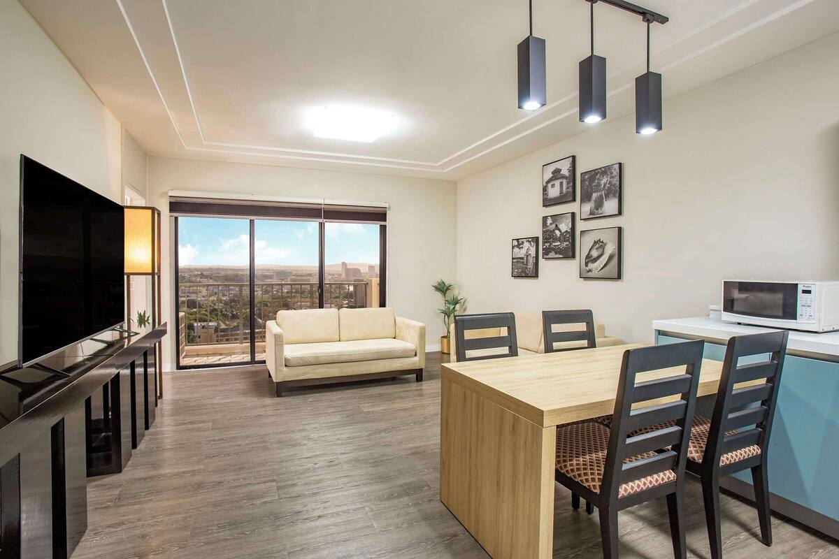 A modern living room with a flat-screen TV, a sofa, a dining table with chairs, and a kitchen area with a microwave.