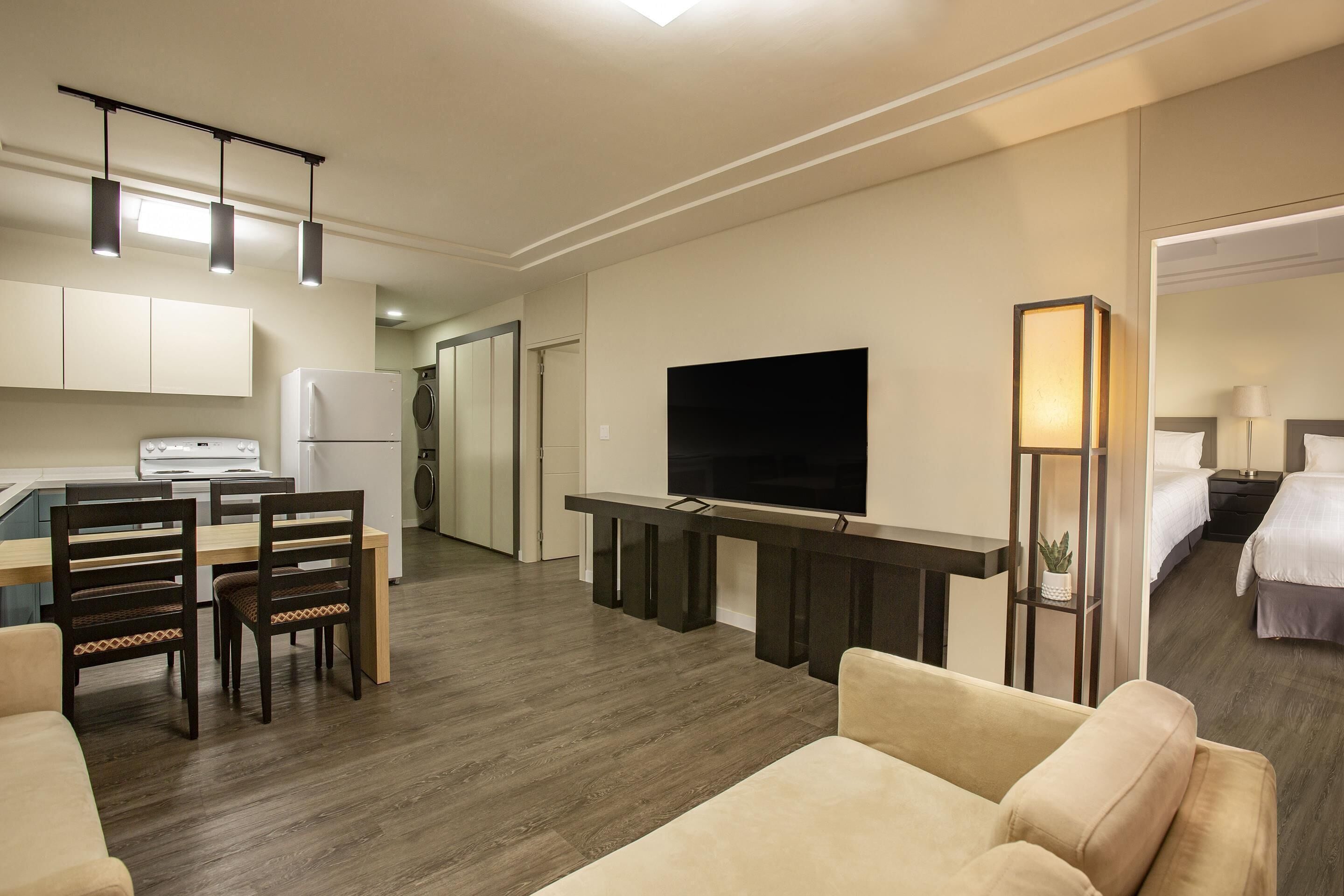 A modern hotel room with a dining area, a flat-screen TV, a sofa, and a bedside table with a lamp.