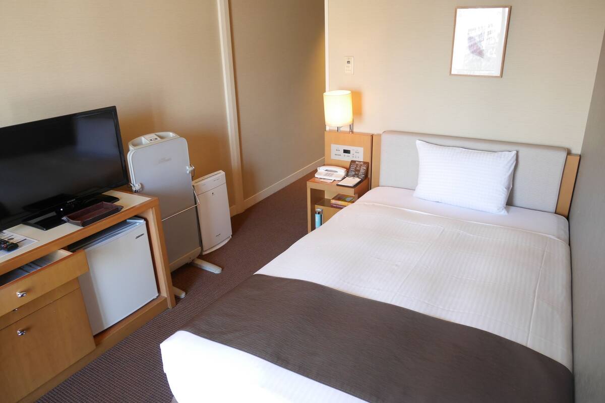 Een hotelkamer met een bed, een bureau met televisie, een koelkast, een nachtkastje met lamp en een wandklok.