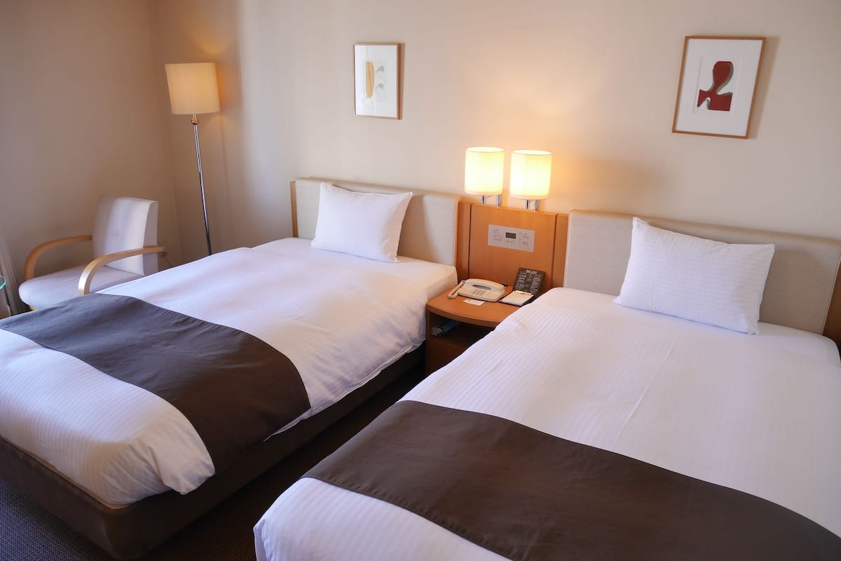 Een hotelkamer met twee bedden, een nachtkastje met een telefoon, een lamp en twee ingelijste foto's aan de muur.
