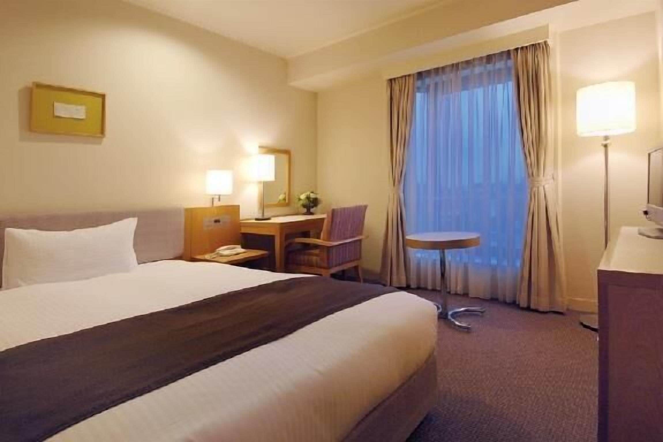 Ett hotellrum med en stor säng, ett skrivbord, en stol, ett litet bord och ett fönster med gardiner.
