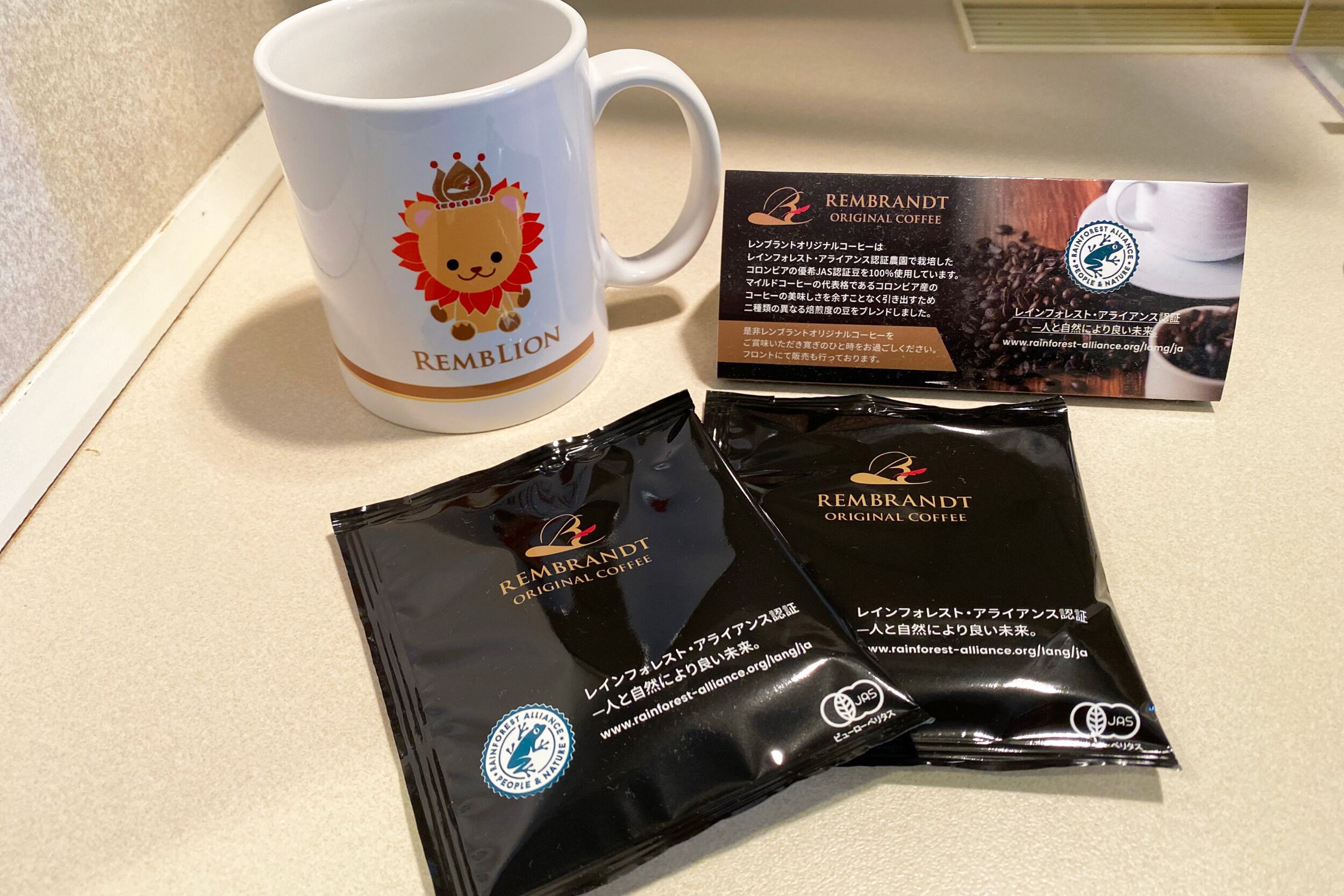 Una taza blanca con diseño de león y la palabra 'REMBRANDT', dos sobres de café negro y una bolsa de café con texto y un logotipo.
