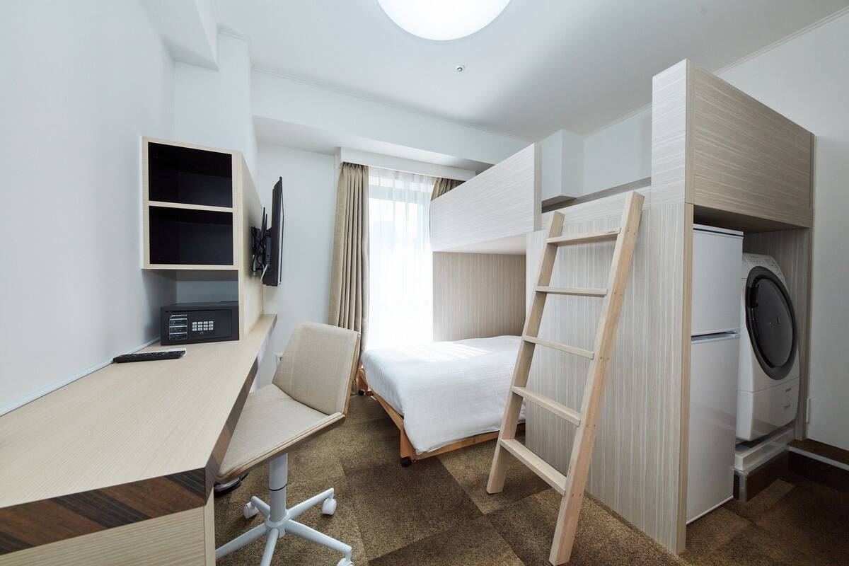 Een compacte hotelkamer met een stapelbed, een bureau met stoel, een wasmachine en een kleine televisie.