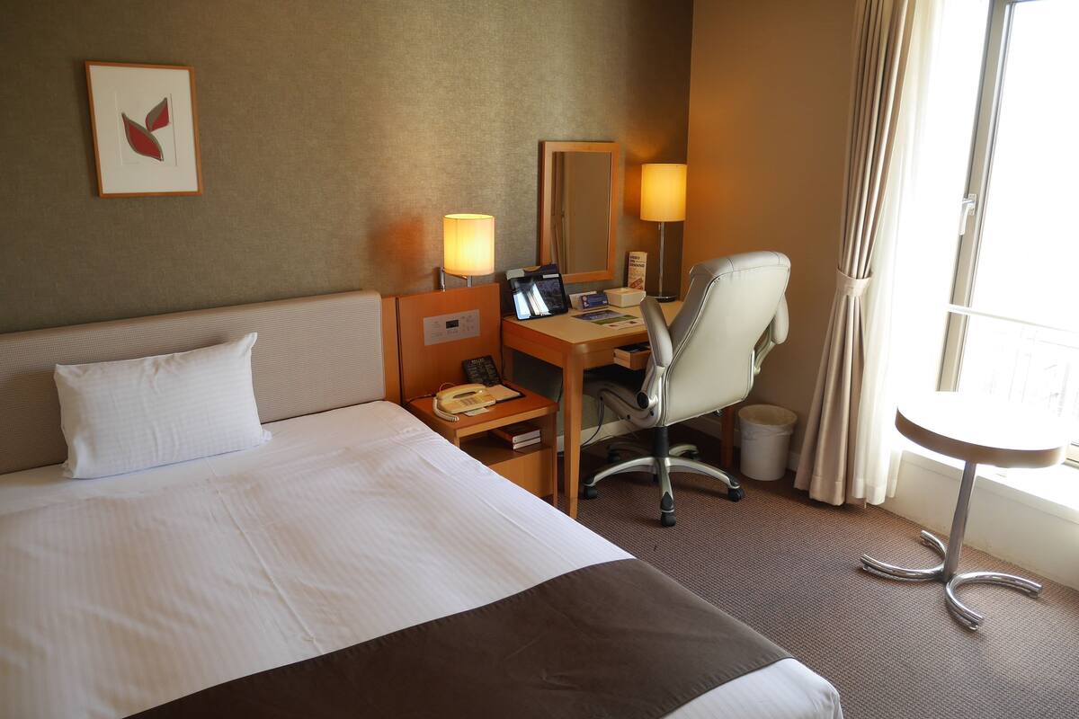 Een hotelkamer met een bed, bureau, stoel, lamp en een raam met gordijnen.