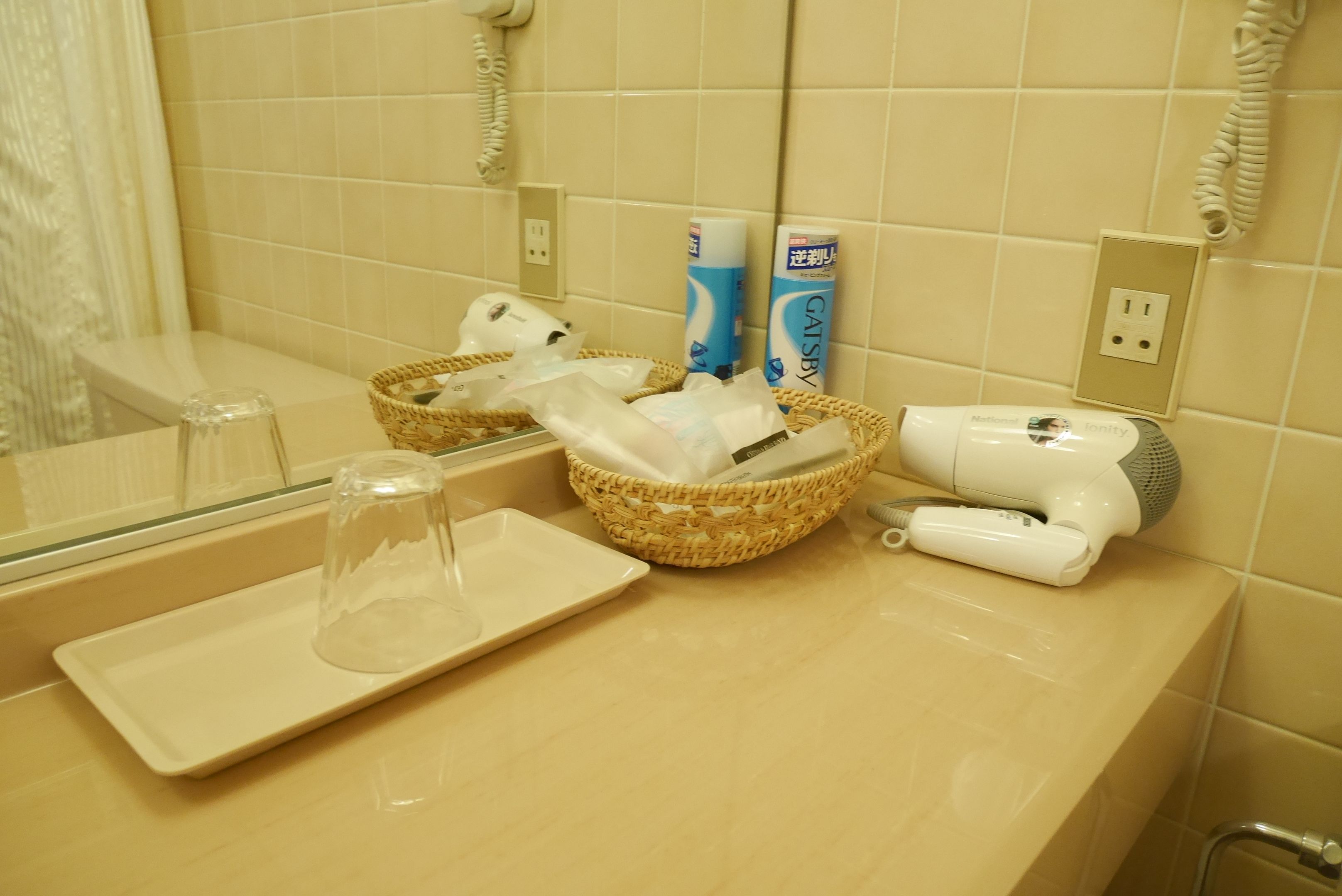 A bathroom with a mirror, a hairdryer, a basket with toiletries, and a glass on a tray.