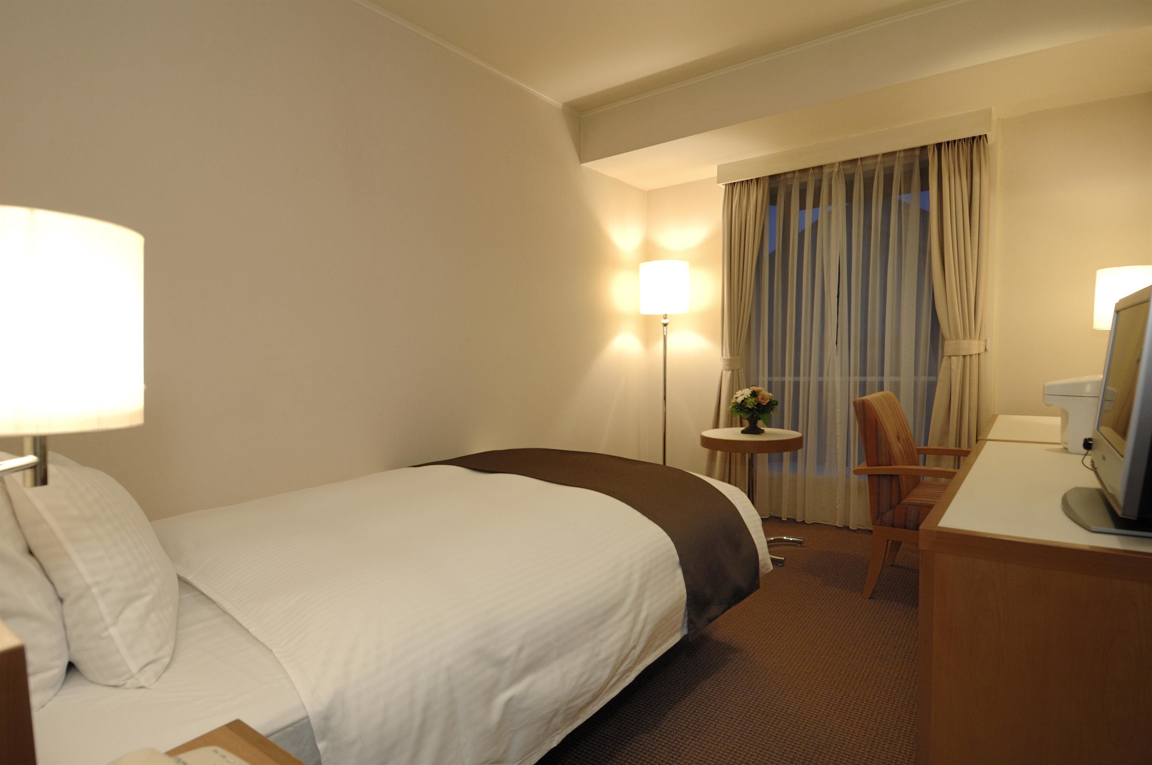 A hotel room with a bed, a desk with a computer, a chair, a lamp, and a window with curtains.
