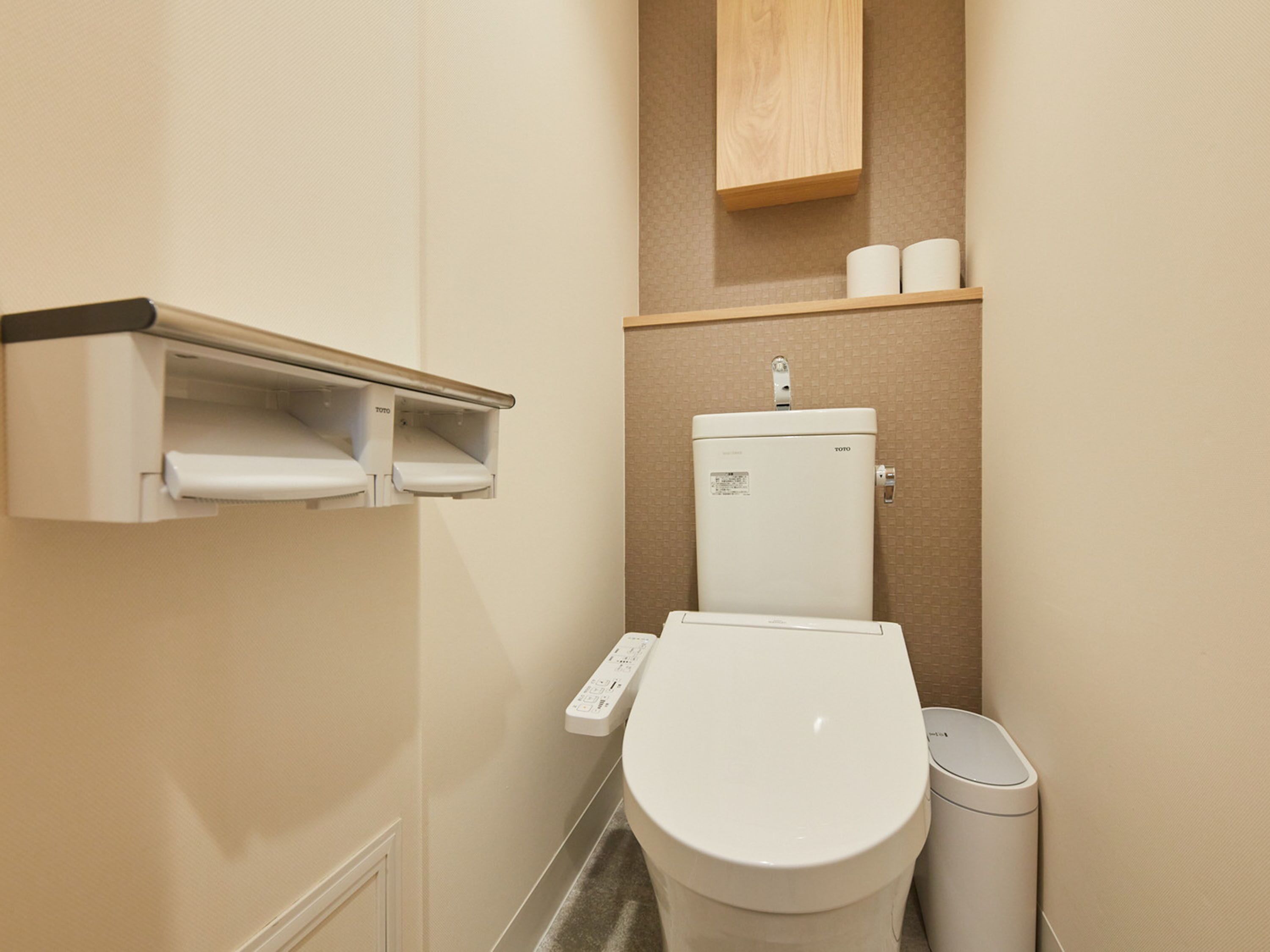 A compact bathroom with a toilet, a trash bin, a shelf with toilet paper, and a paper towel dispenser.