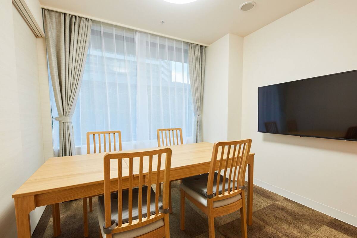 Een kamer met een houten eettafel, vier stoelen, een groot raam met dunne gordijnen en een flatscreen televisie aan de muur.