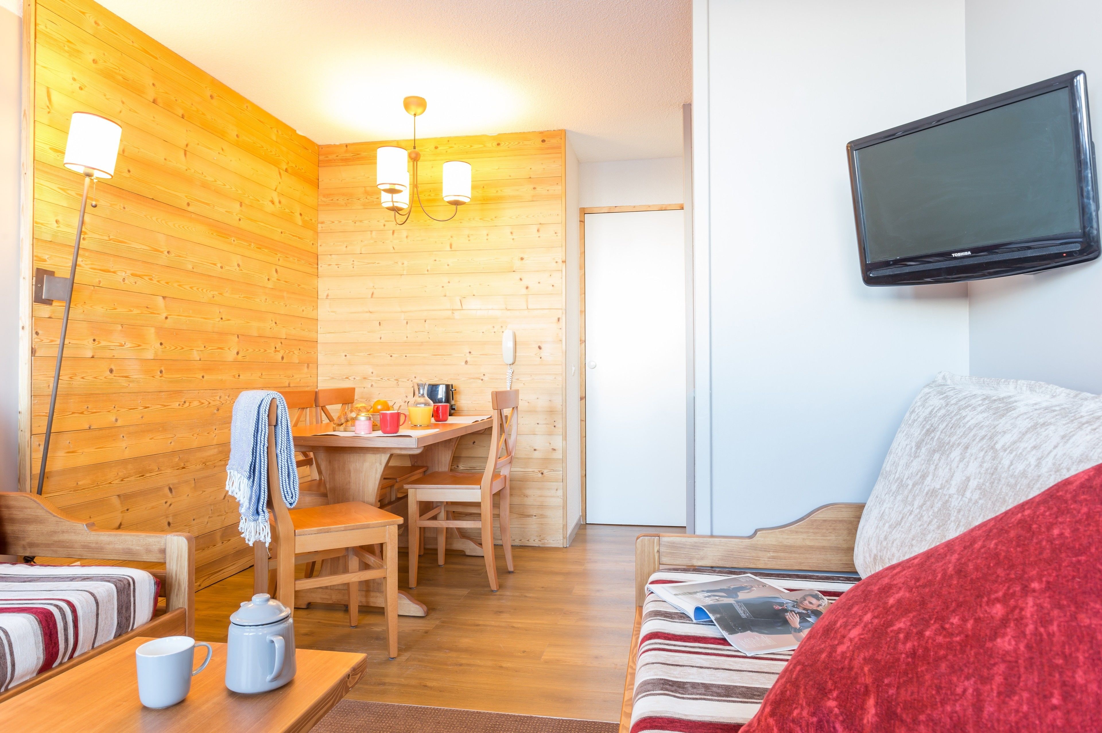 A cozy room with wooden walls, a flat-screen TV mounted on the wall, a dining area with a table and chairs, and a bed with a striped blanket.