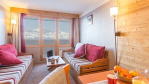 A cozy room with two beds, a table with a bowl of fruit, and a view of snow-capped mountains through large windows.