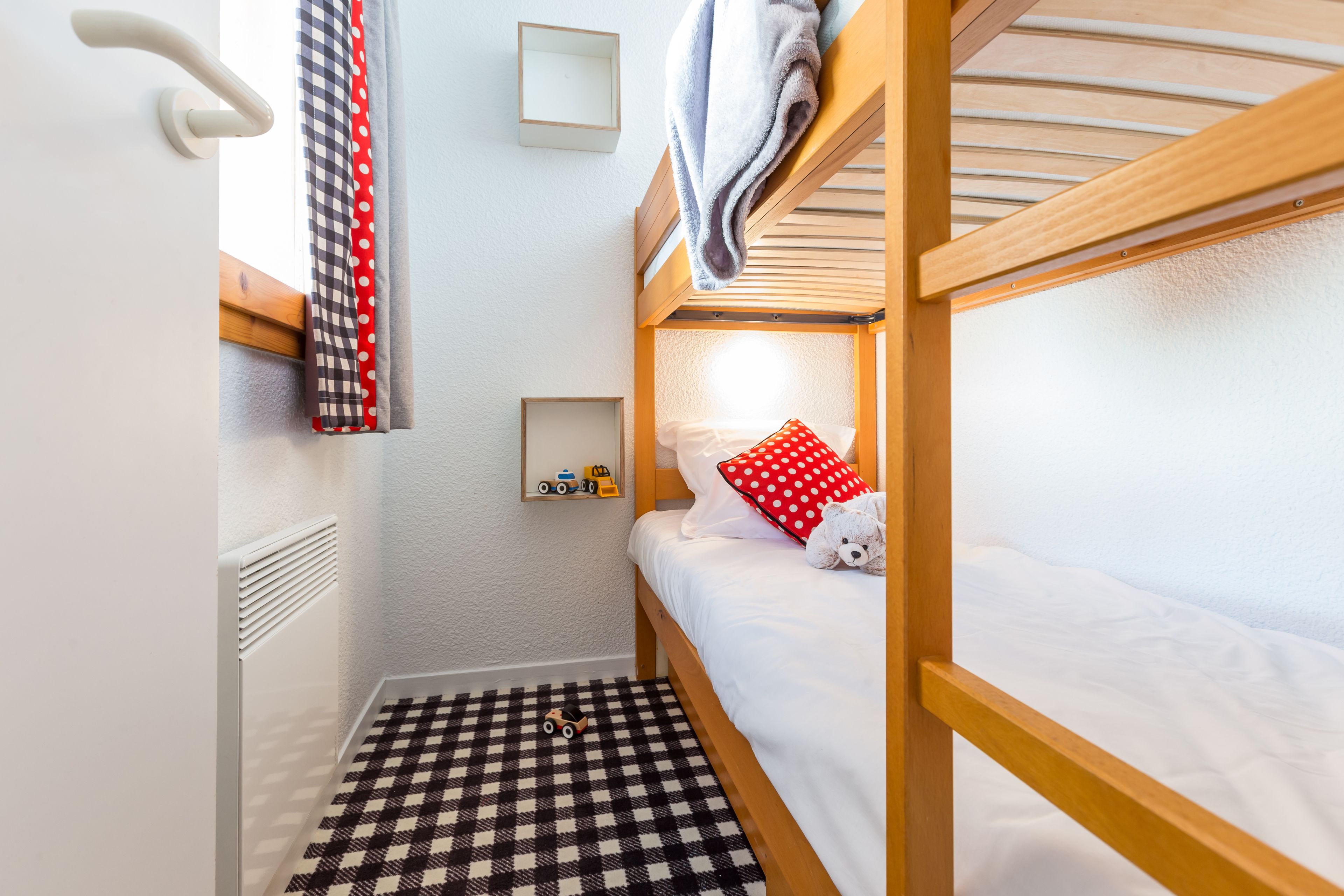 Une petite pièce accueillante, avec un lit superposé, un sol à carreaux, un radiateur, une étagère remplie de jouets et une fenêtre avec des rideaux.