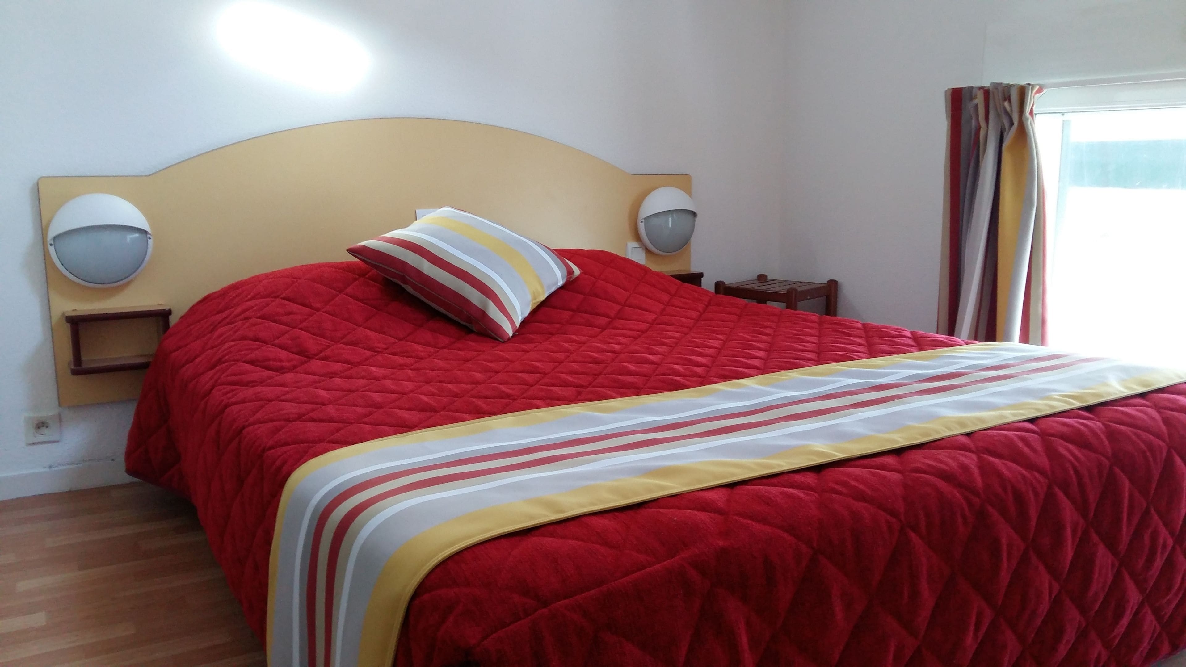 A bed with a red quilted bedspread and a striped pillow, a wooden headboard, two wall-mounted lamps, and a window with curtains.