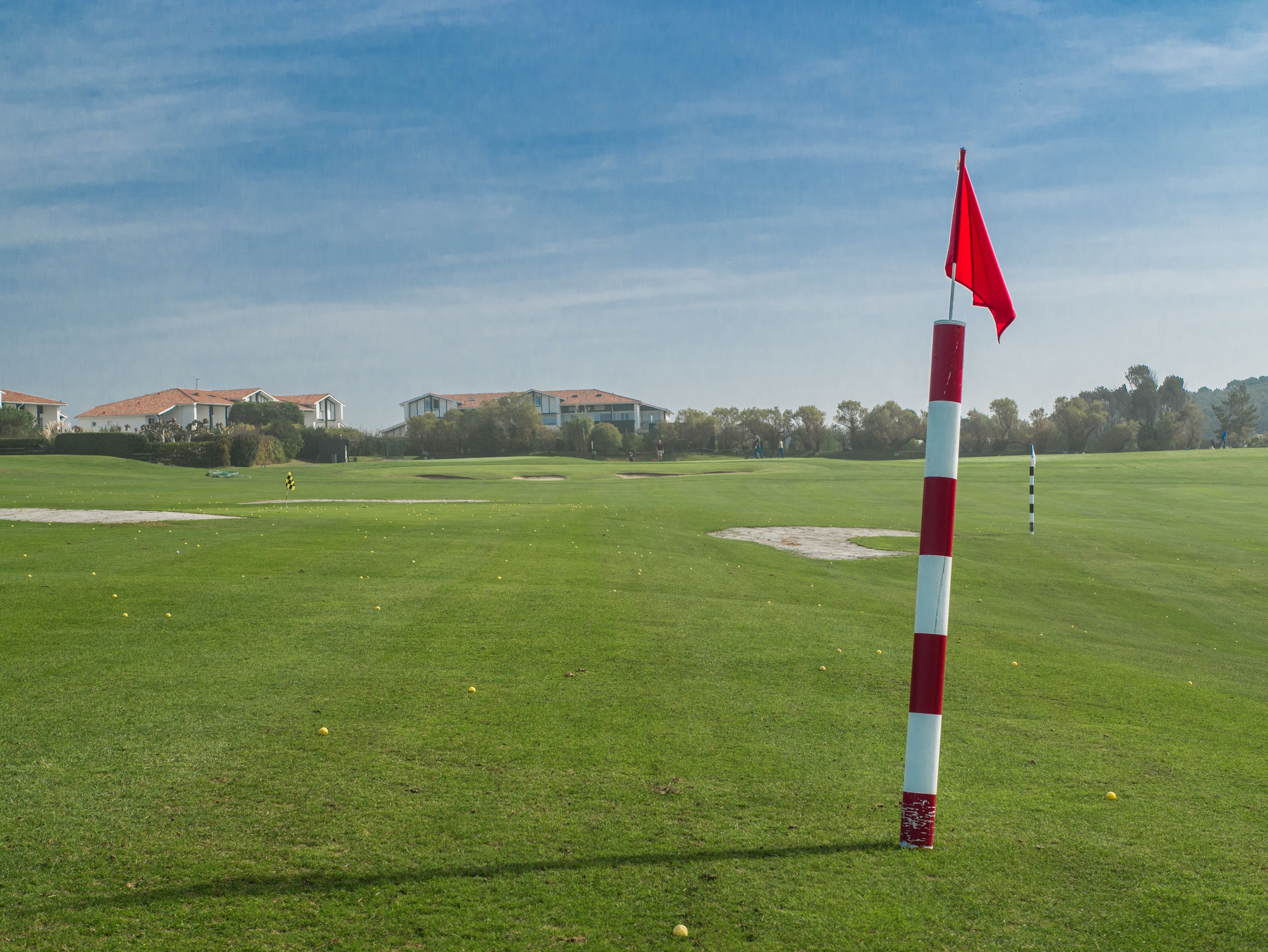A golf course with a red and white flag marker, sand bunkers, and houses in the background.