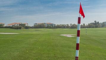 Campo de golf con un marcador de bandera rojo y blanco, búnkeres de arena y casas al fondo.