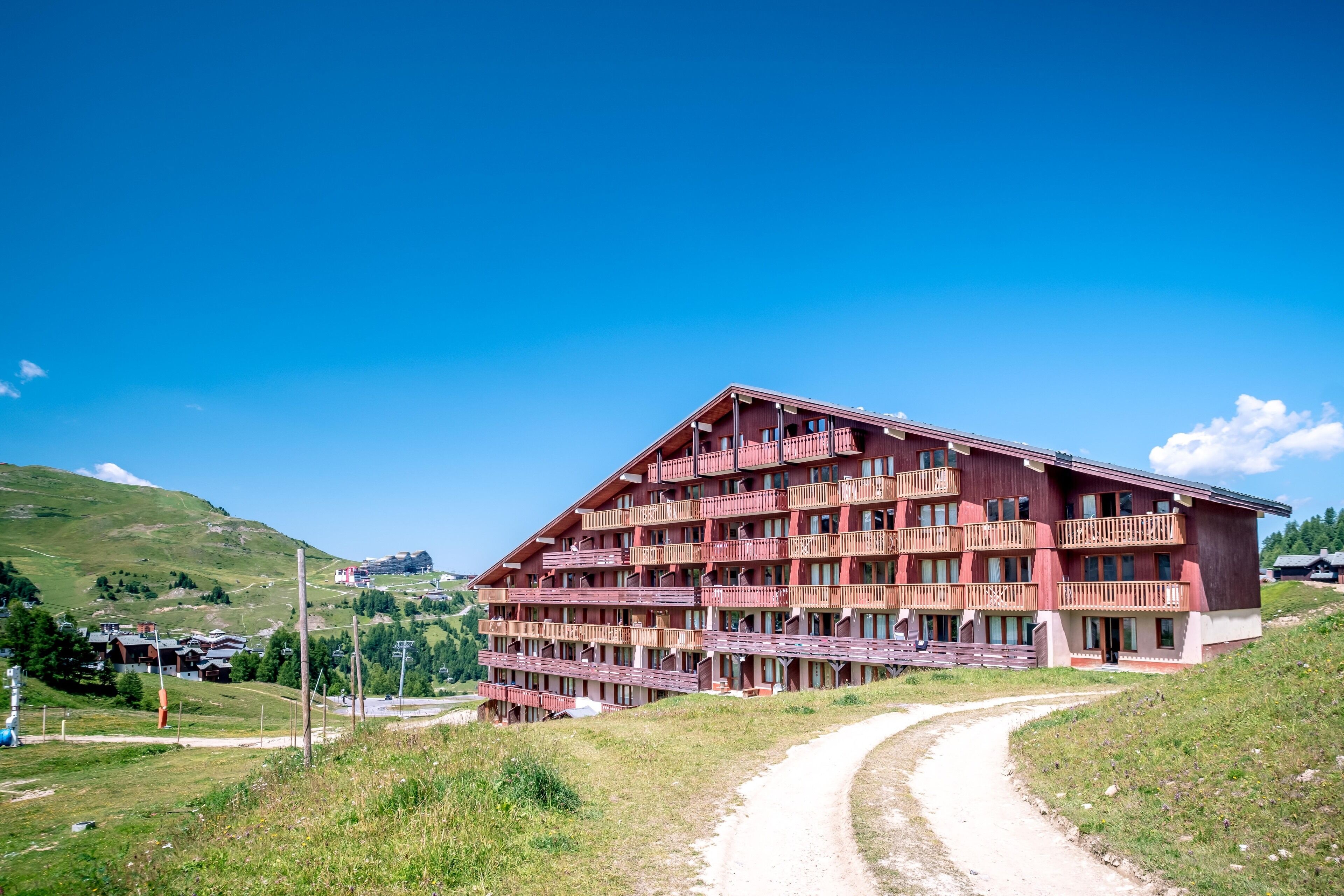 Un hotel de varios pisos con balcones, ubicado en una ladera con un camino de tierra que lo lleva a él.