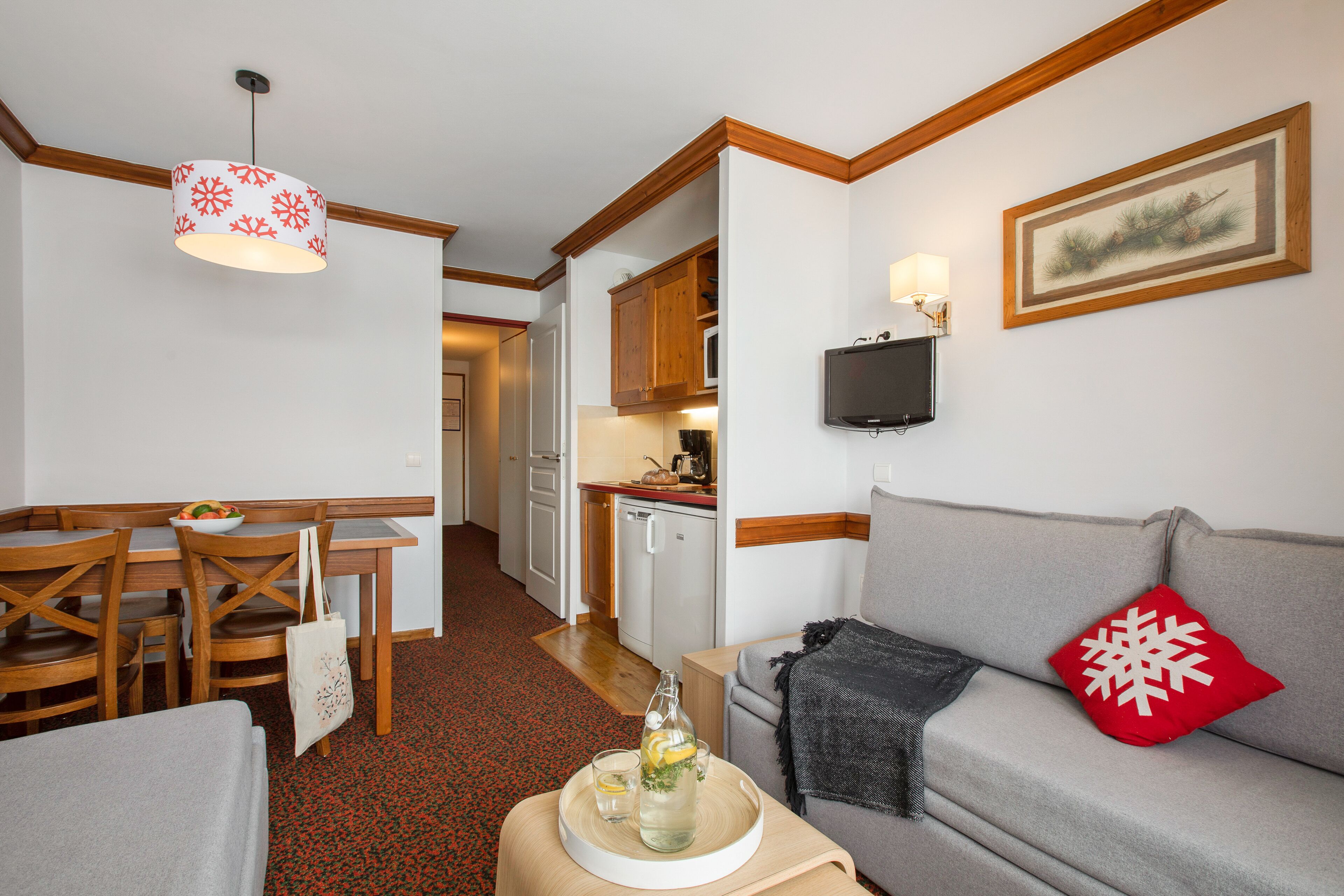 Un espace de vie compact comprenant un coin repas, une kitchenette et un canapé gris agrémenté d’un coussin rouge.