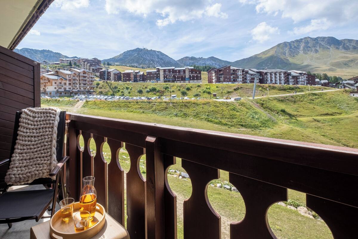 A balcony with a wooden railing, a chair, a table with a glass of beer, and a view of a mountainous town with buildings.