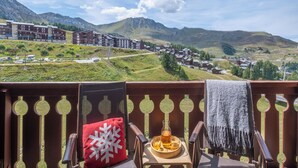 A balcony with a view of a mountainous town, a wooden chair, a red pillow, and a tray with a glass and a bottle.