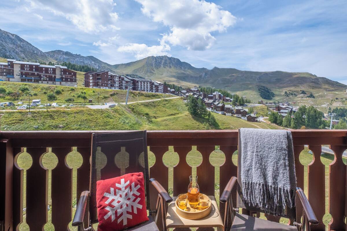 A balcony with a view of a mountainous town, a wooden chair, a red pillow, and a tray with a glass and a bottle.