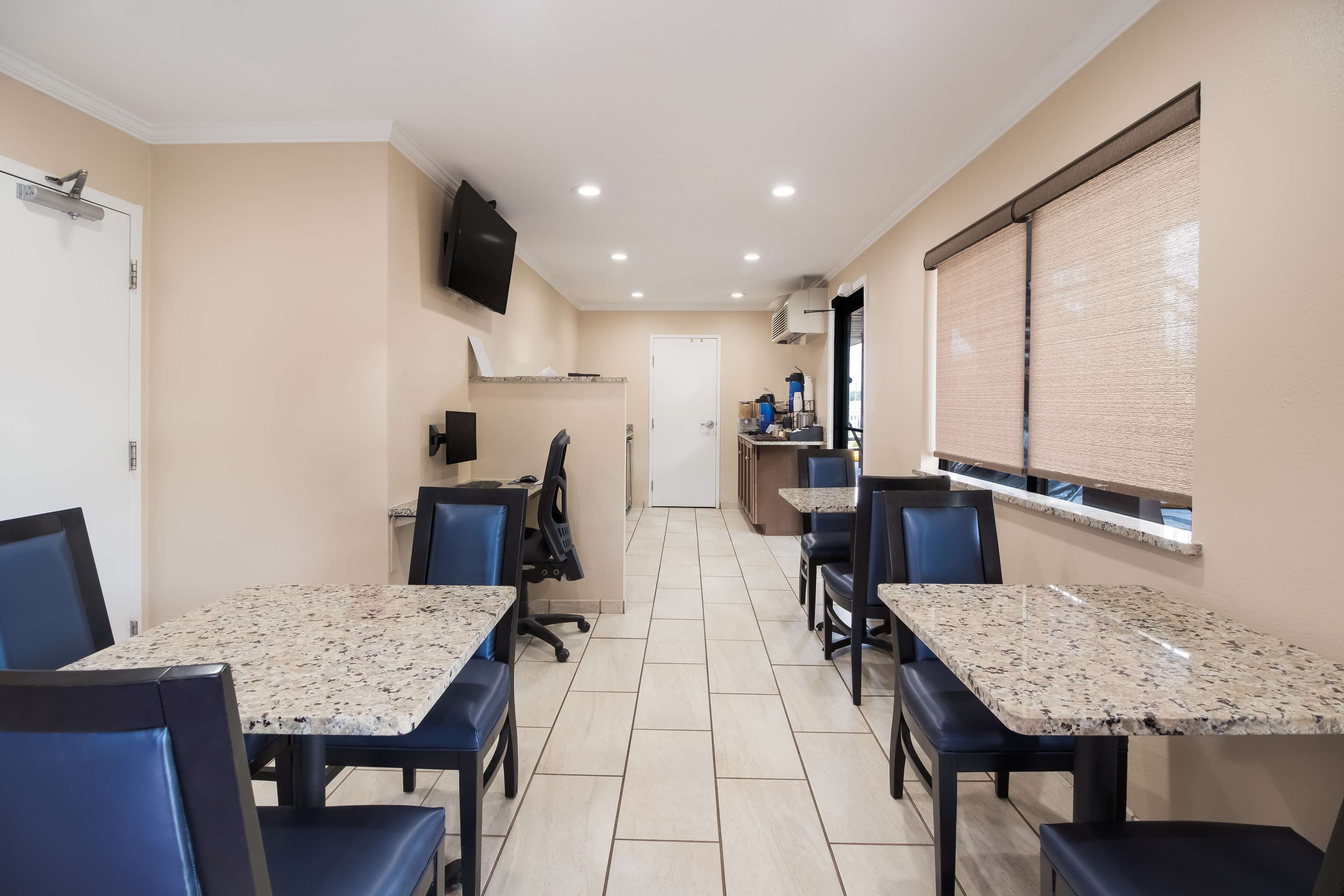 A hotel dining area with granite tables and blue chairs, a flat-screen TV on the wall, and a window with blinds.