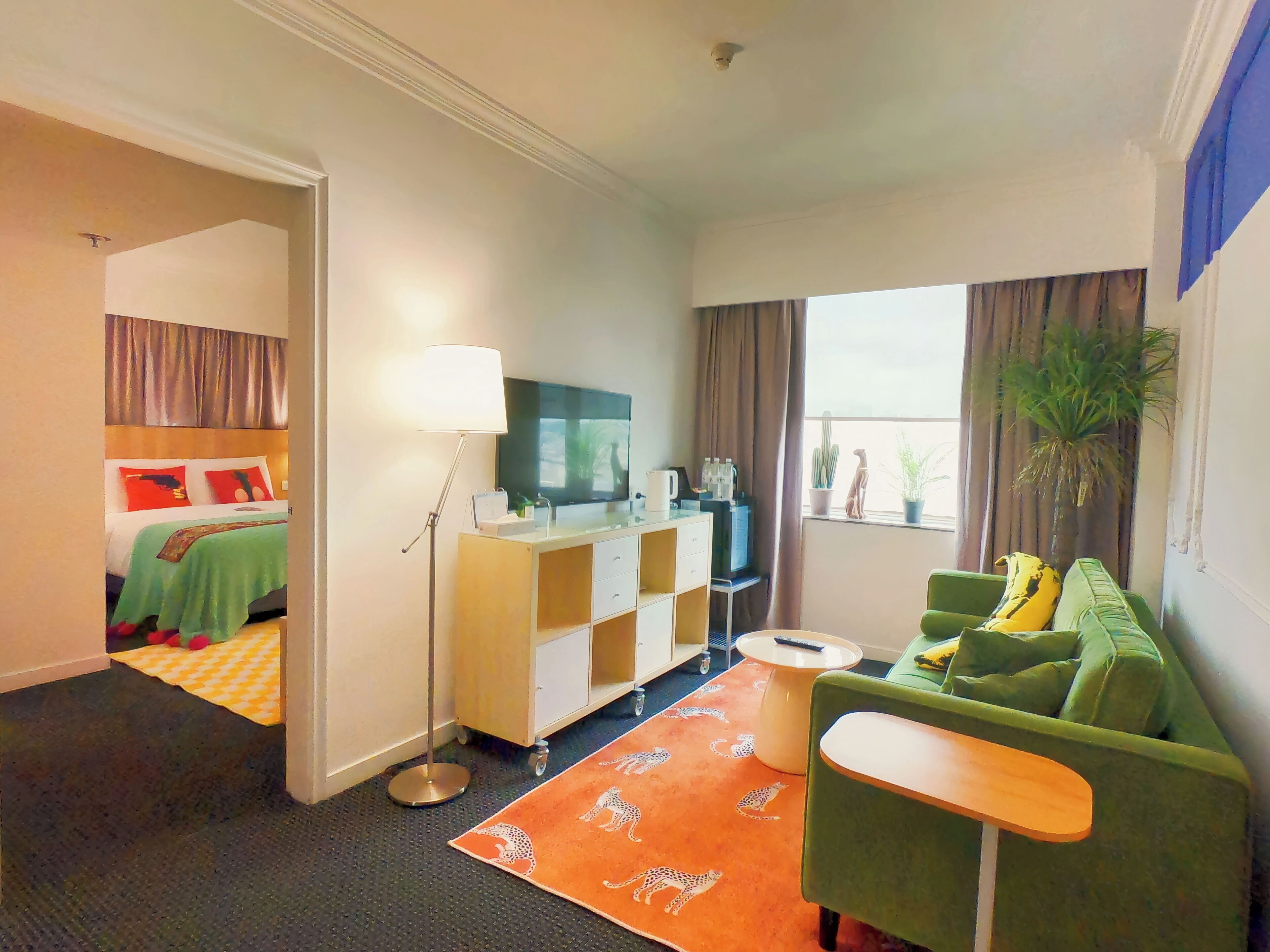 A hotel room with a bed, a desk, a green sofa, a small table, a lamp, a TV, and a window with curtains.