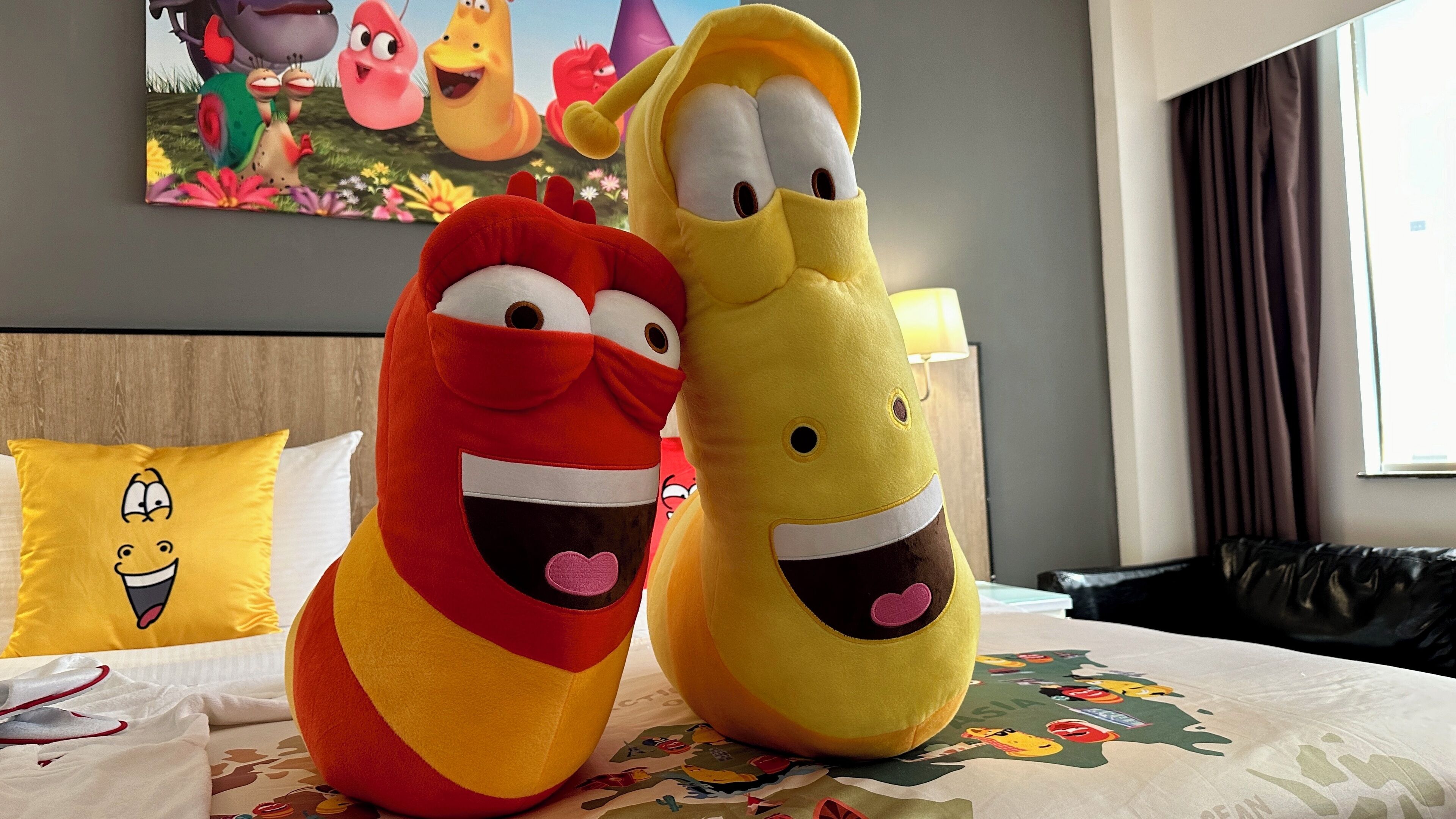 Two plush toys on a bed with a pillow featuring a cartoon character, a lamp, and a window with curtains.