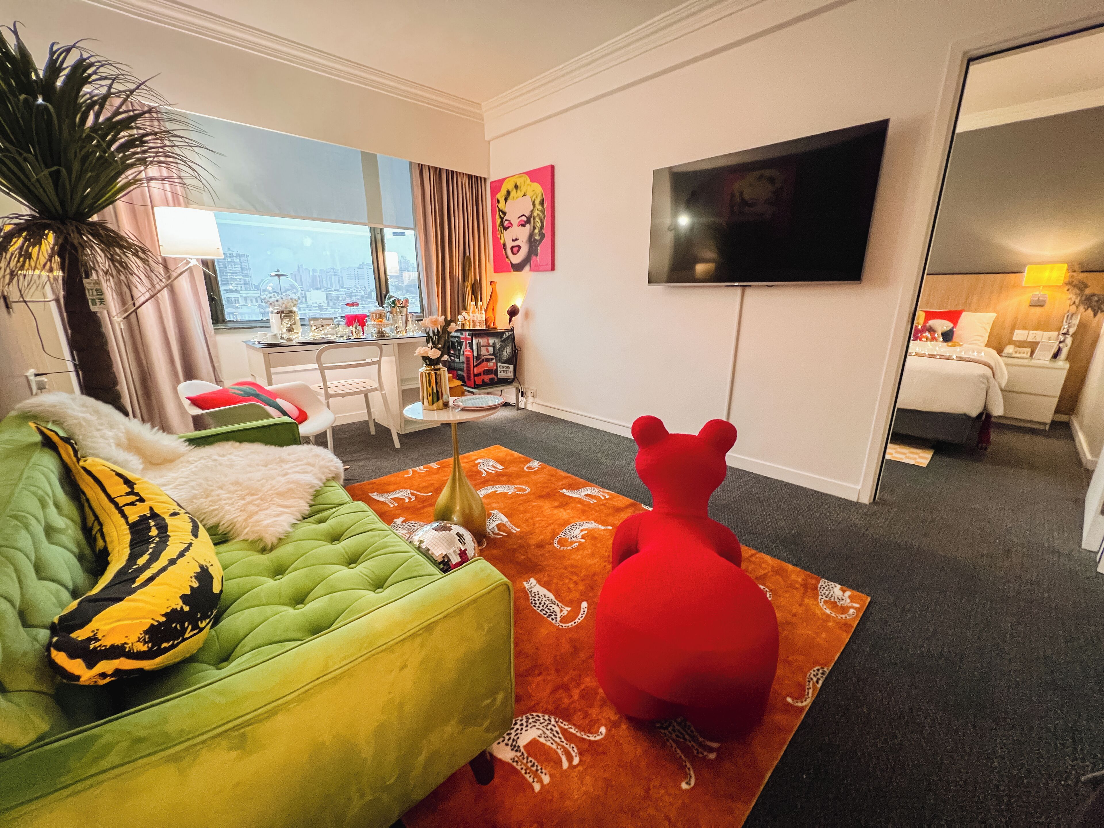 A modern living room with a green sofa, a red bear-shaped rug, and a city view through large windows.
