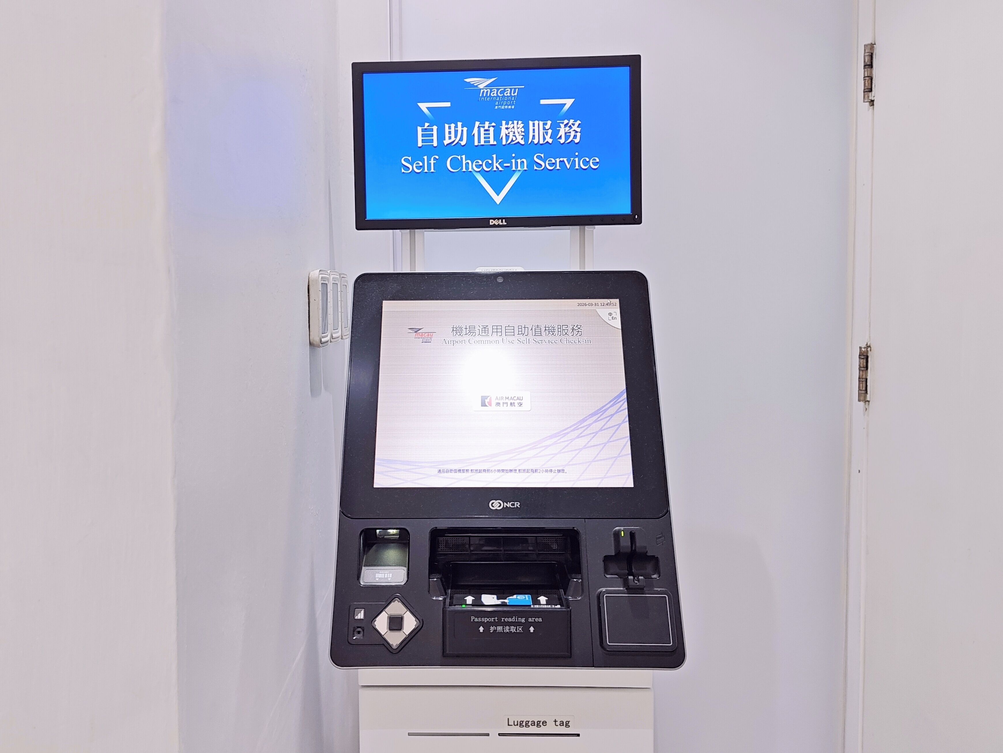 A self-service check-in kiosk with a monitor displaying instructions in multiple languages, a passport reading area, and a luggage tag dispenser.