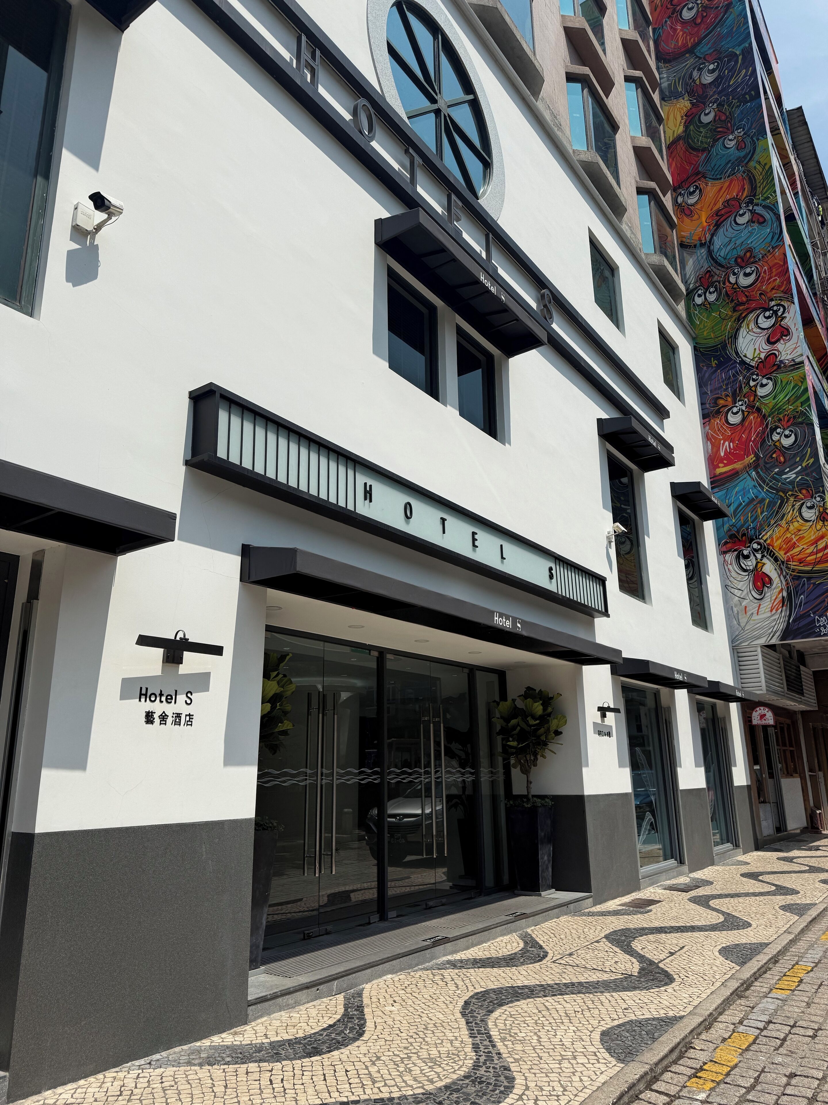 A modern hotel with a prominent 'HOTEL S' sign, large windows, and a colorful mural on the adjacent building.