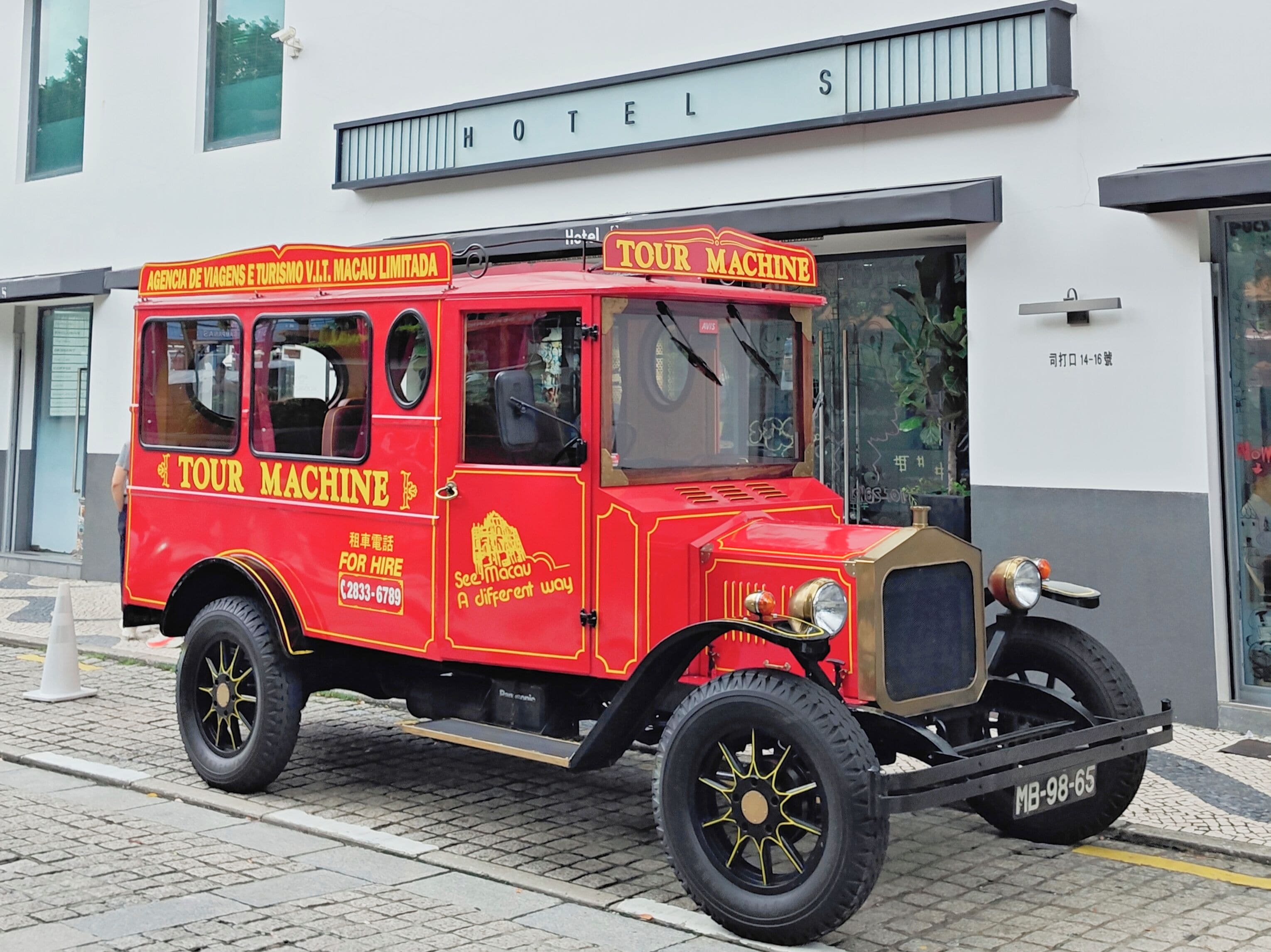A red vintage tour bus parked in front of a hotel with a sign that reads 'TOUR MACHINE'.
