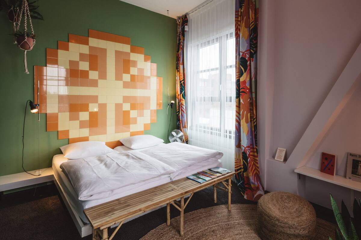 A bedroom with a bed, a bench, a wall-mounted lamp, a fan, and a window with curtains.