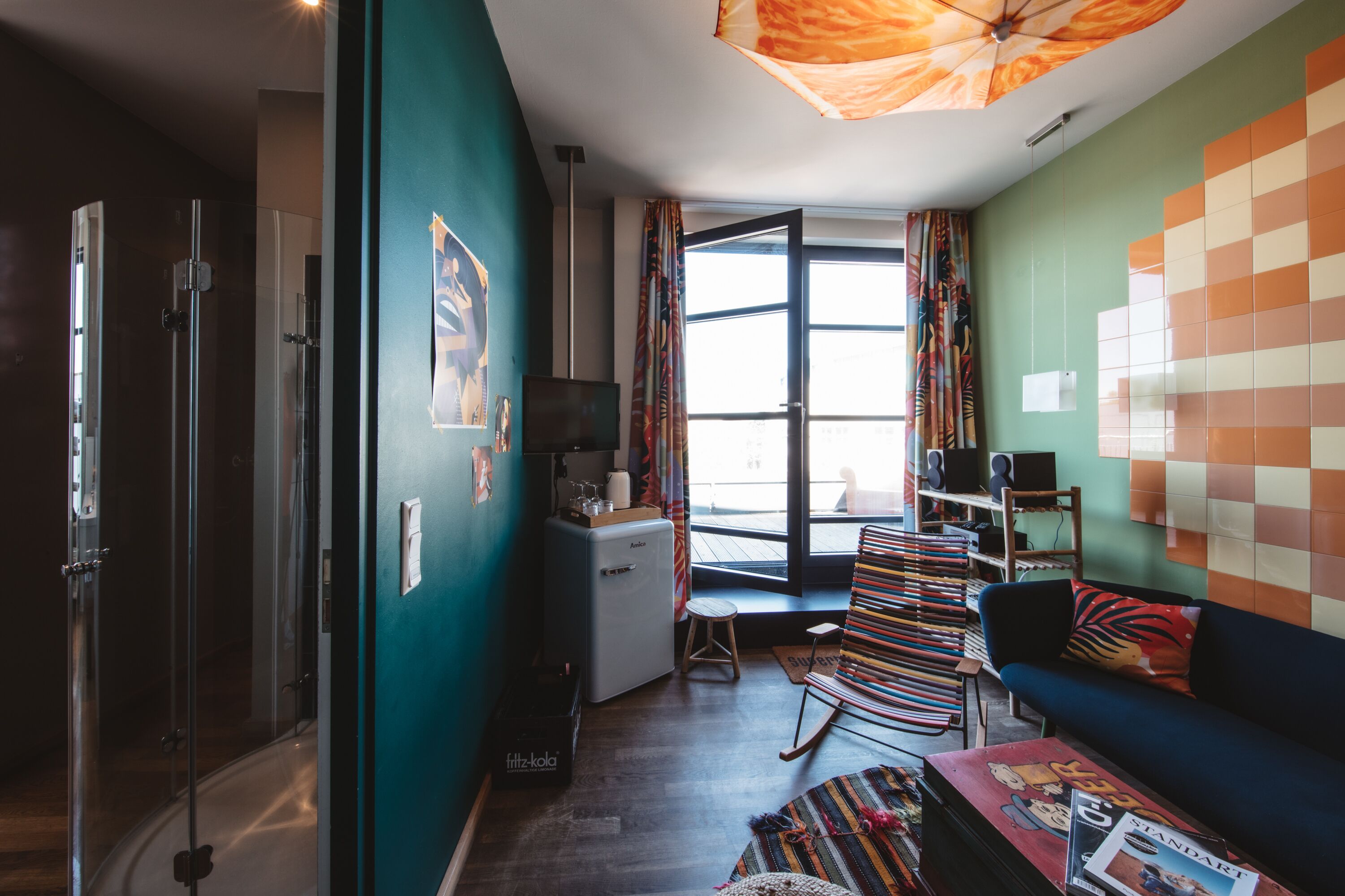 A compact living space with a kitchenette, a sofa, a colorful chair, and a glass shower enclosure.