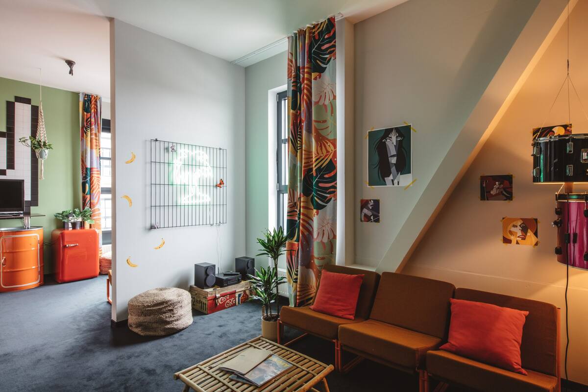 A living room with a sofa, a coffee table, and a TV. There are plants, a curtain, and a wall with pictures.