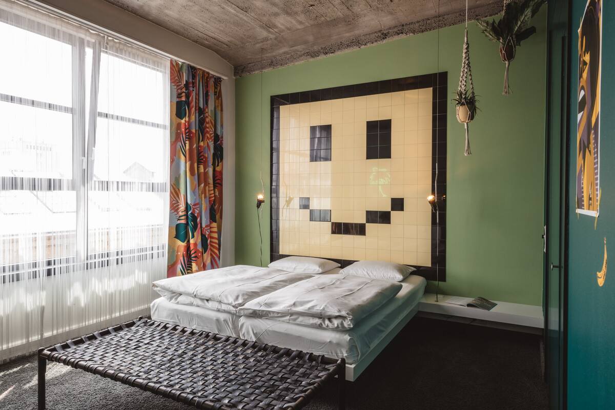 A room with a bed, a bench, a window with curtains, and a wall with a geometric pattern.