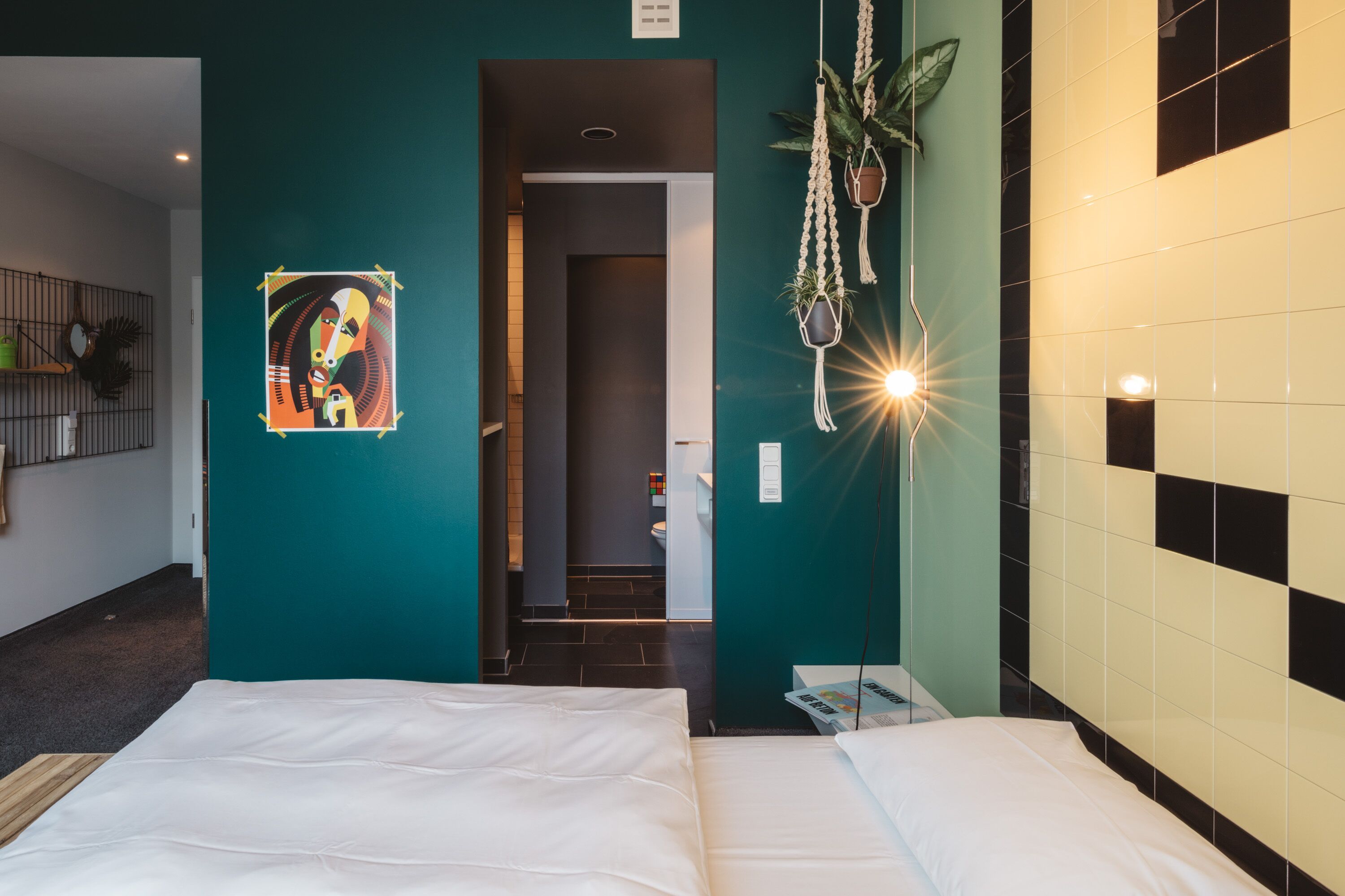 A hotel room with a teal accent wall, a bed with white bedding, a door leading to a bathroom, and a hanging plant.