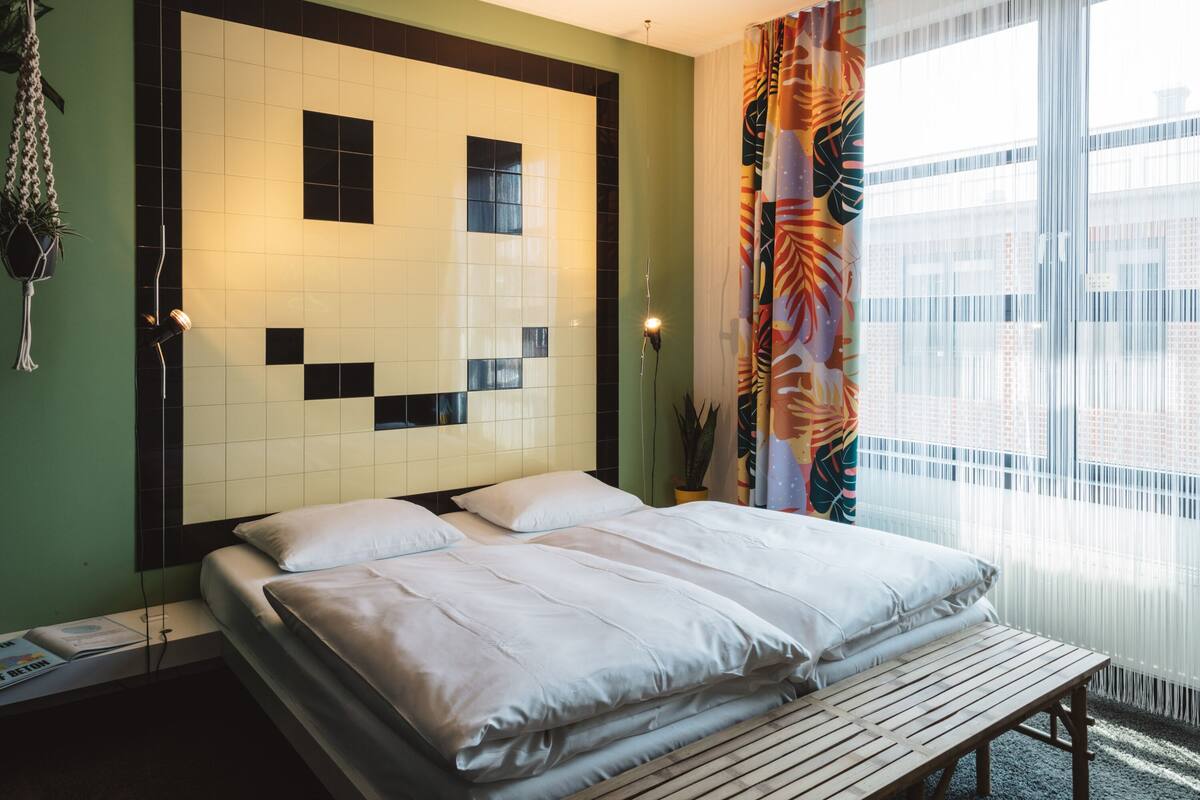 A hotel room with a bed, a wooden bench, a wall with a geometric pattern, and a window with curtains.