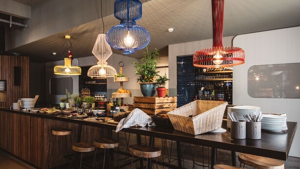 A modern bar area with multiple hanging light fixtures, a bar counter with stools, and a variety of bar tools and bottles. - Superbude Altona Paradise (Hamburg)